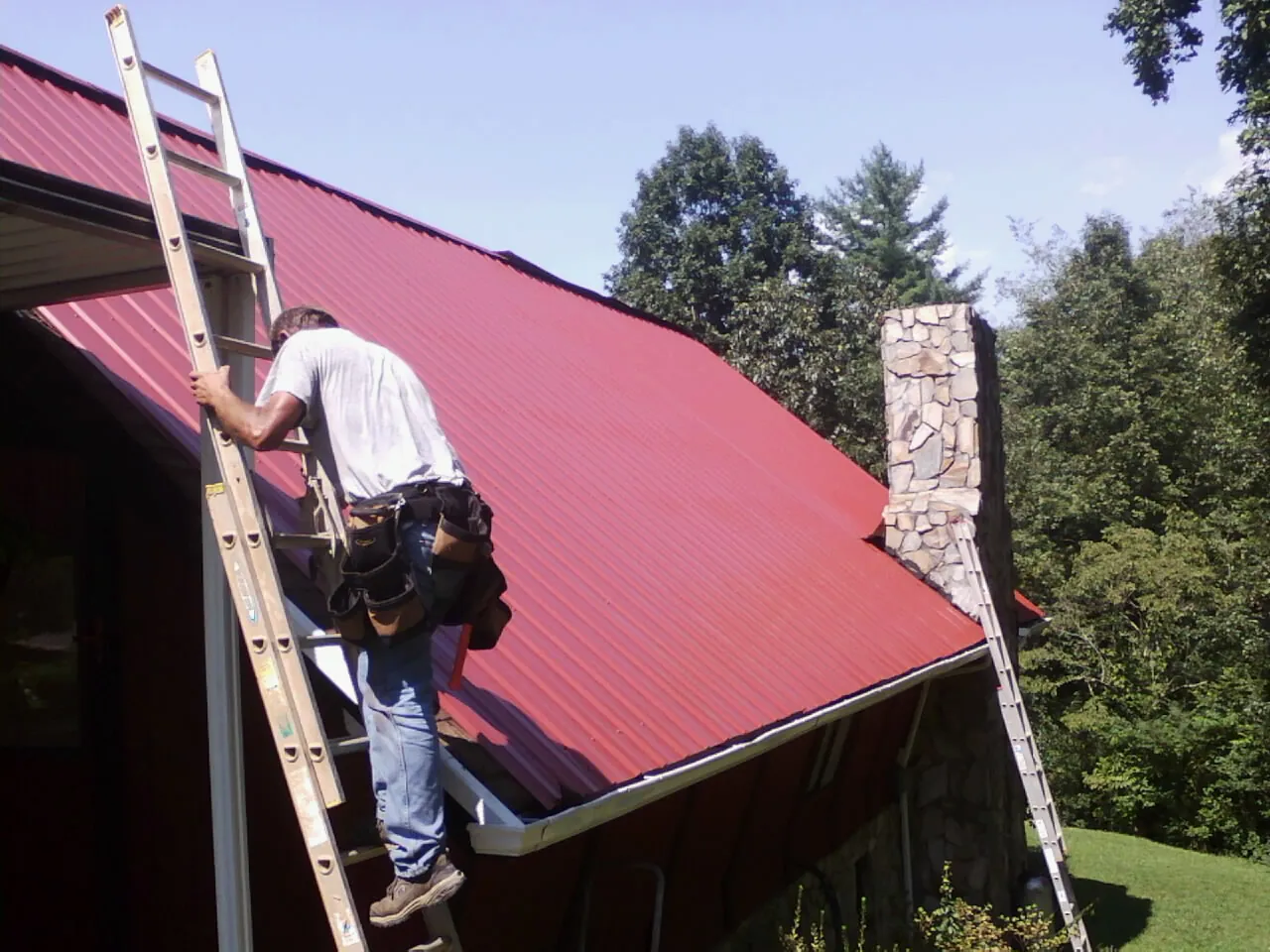 Roofer on a ladder beside a red metal roof and stone chimney — Soffit & Fascia Repair by Roofing Brothers in Eighty Four